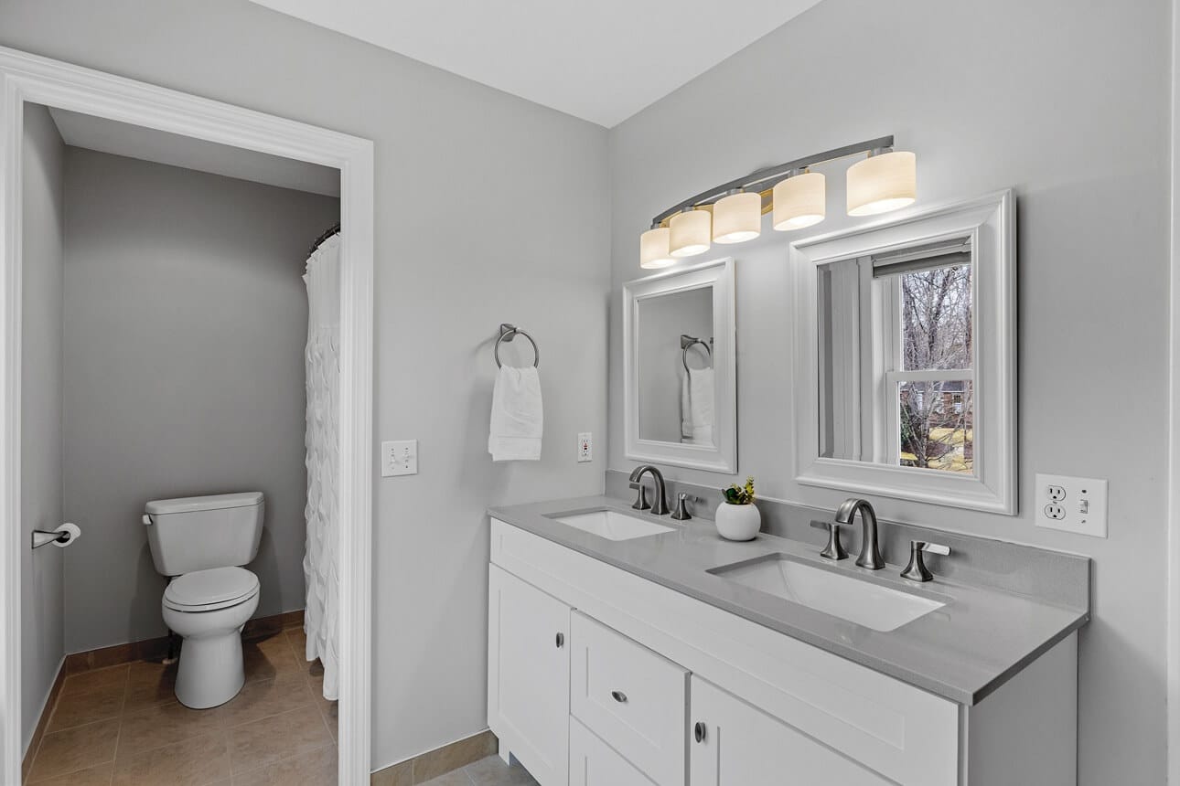 Bathroom Remodeling Services in Fredericksburg Texas​ Modern bathroom with dual sinks, gray walls, and natural light, featuring a white toilet and a shower curtain in the background, highlighting stylish design and functional layout for remodeling services in Fredericksburg, Texas.