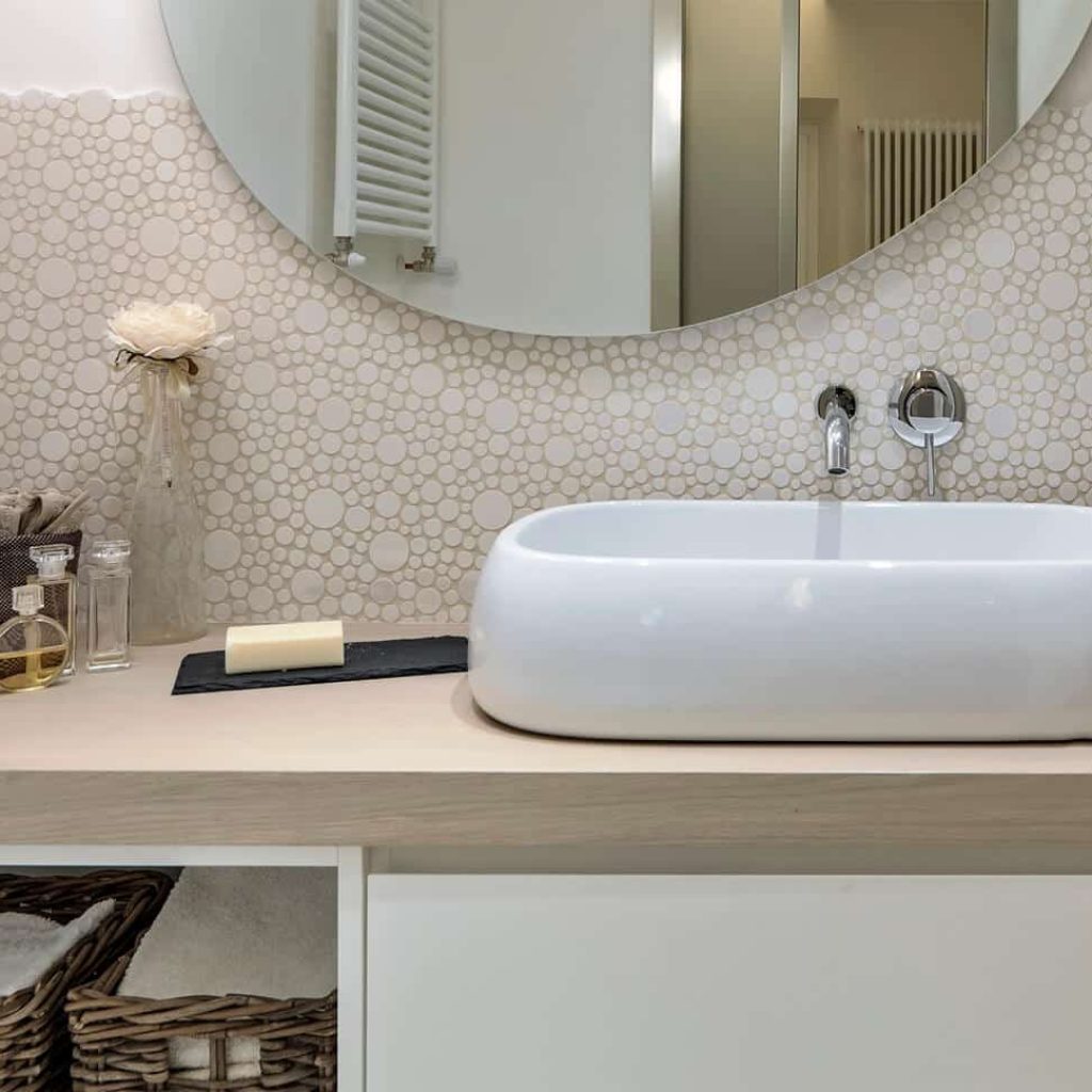 Modern bathroom design featuring a white vessel sink on a wooden countertop, surrounded by decorative pebble-patterned backsplash, elegant accessories, and a round mirror, showcasing a stylish renovation ideal for Kerrville homeowners.